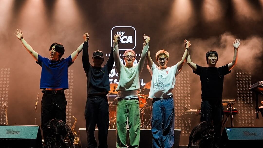 Members of Wave to Earth stand on stage at Java Jazz Festival 2026, holding hands and raising their arms to acknowledge the audience beneath bright concert spotlights and atmospheric stage smoke.